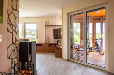 [Bungalov 4 kk s terasou, obývák s krbovými kamny a kamenným obložením, průhled do cvičící místnosti a do zahrady.]