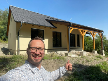 [Rodinný dům Haas, dřevostavba, individuální bungalov 3+kk. Pohled zvenku na zastřešenou terasu. Menší dům se sedlovou střechou.]